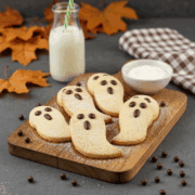 Grafted Pro ghost-shaped Halloween cookies with chocolate eyes on wooden board, milk, and whipped cream, autumn leaves, cozy bakery setting, perfect for fall celebrations and spooky treats.