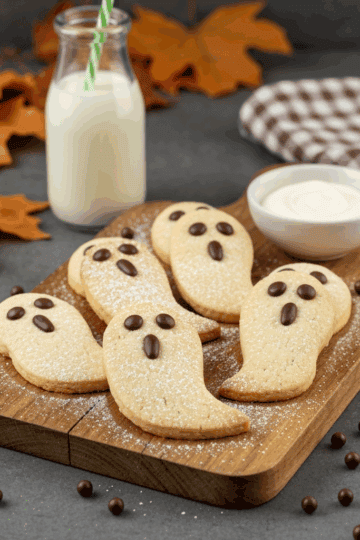 Grafted Pro ghost-shaped Halloween cookies with chocolate eyes on wooden board, milk, and whipped cream, autumn leaves, cozy bakery setting, perfect for fall celebrations and spooky treats.